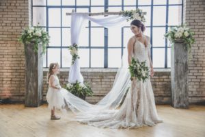 flowergirl holding the veil of a bride in a lace wedding dress while she looks back at her in front of a large window