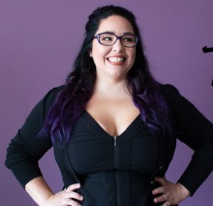 A woman with purple hair and glasses posing for the camera.